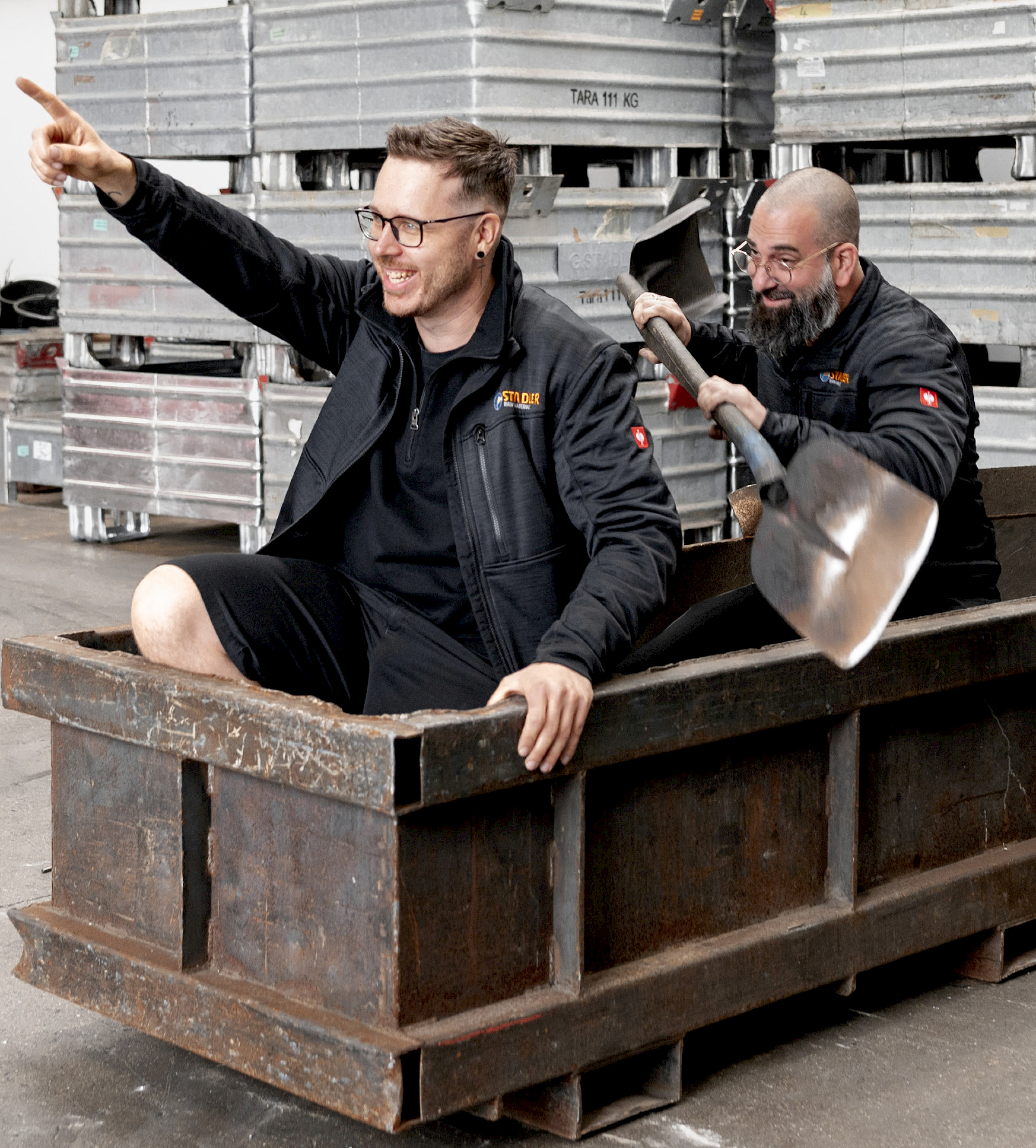 Zwei Männer in Arbeitskleidung sitzen in einem großen, rechteckigen Metallbehälter. Einer zeigt nach vorne, der andere hält eine Schaufel. Im Hintergrund sind gestapelte Metallteile zu sehen.