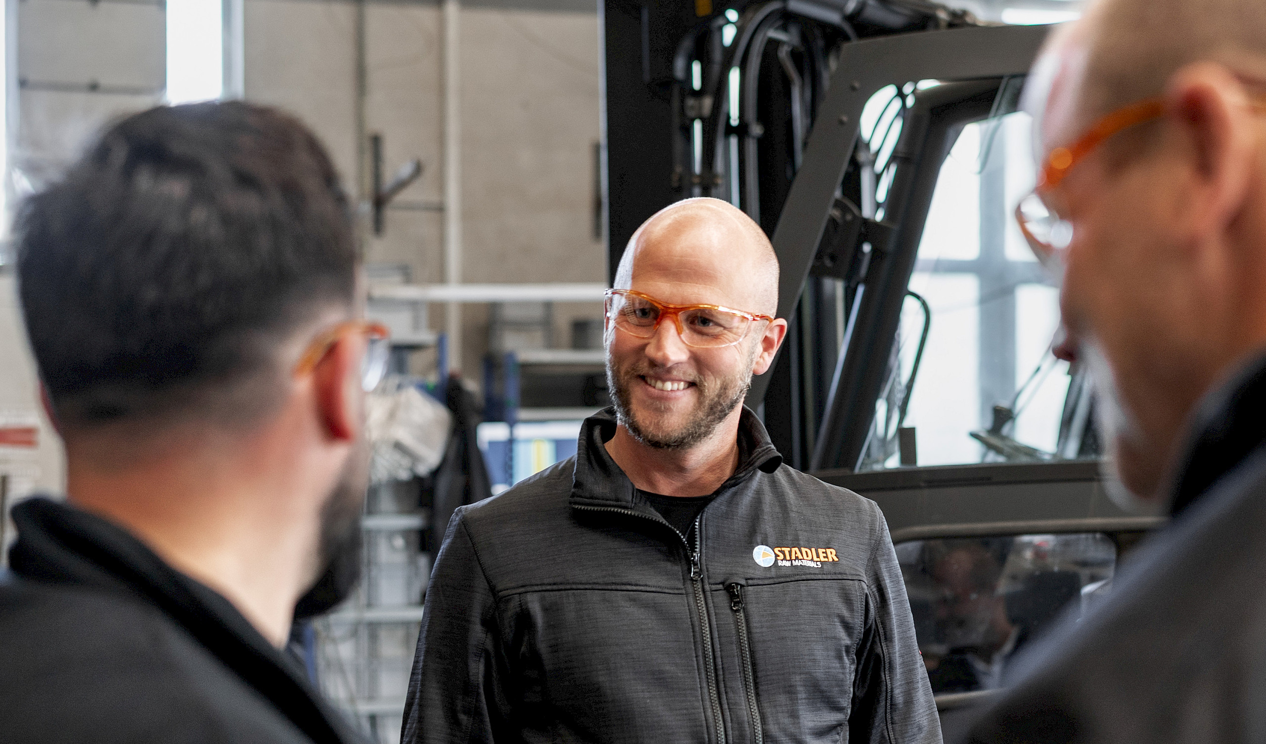 Drei Personen lächeln einander zu. Im Hintergrund ist ein Gabelstapler zu sehen.